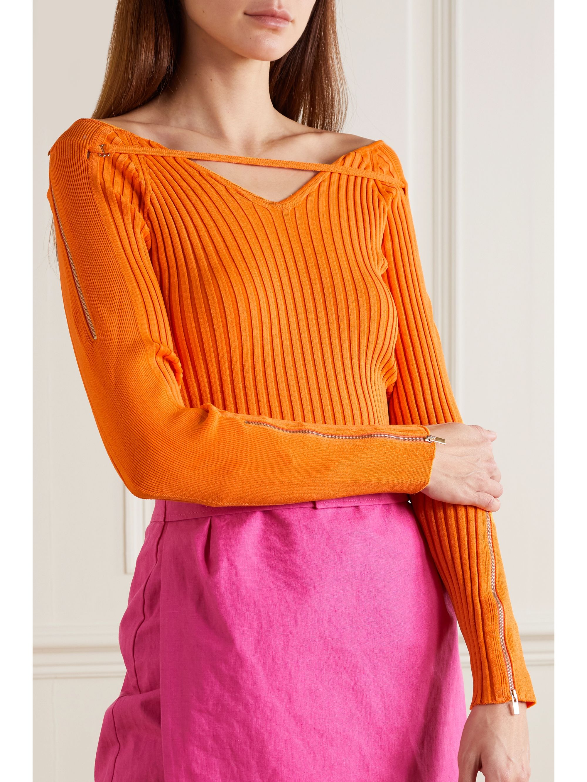 This JACQUEMUS Oro off-the-shoulder cutout ribbed-knit top in orange has a wide v neck which shows off the shoulders. It comes in a rib knit which hugs the body. It has long sleeves with zip details and a signature strap detail at the neck. 