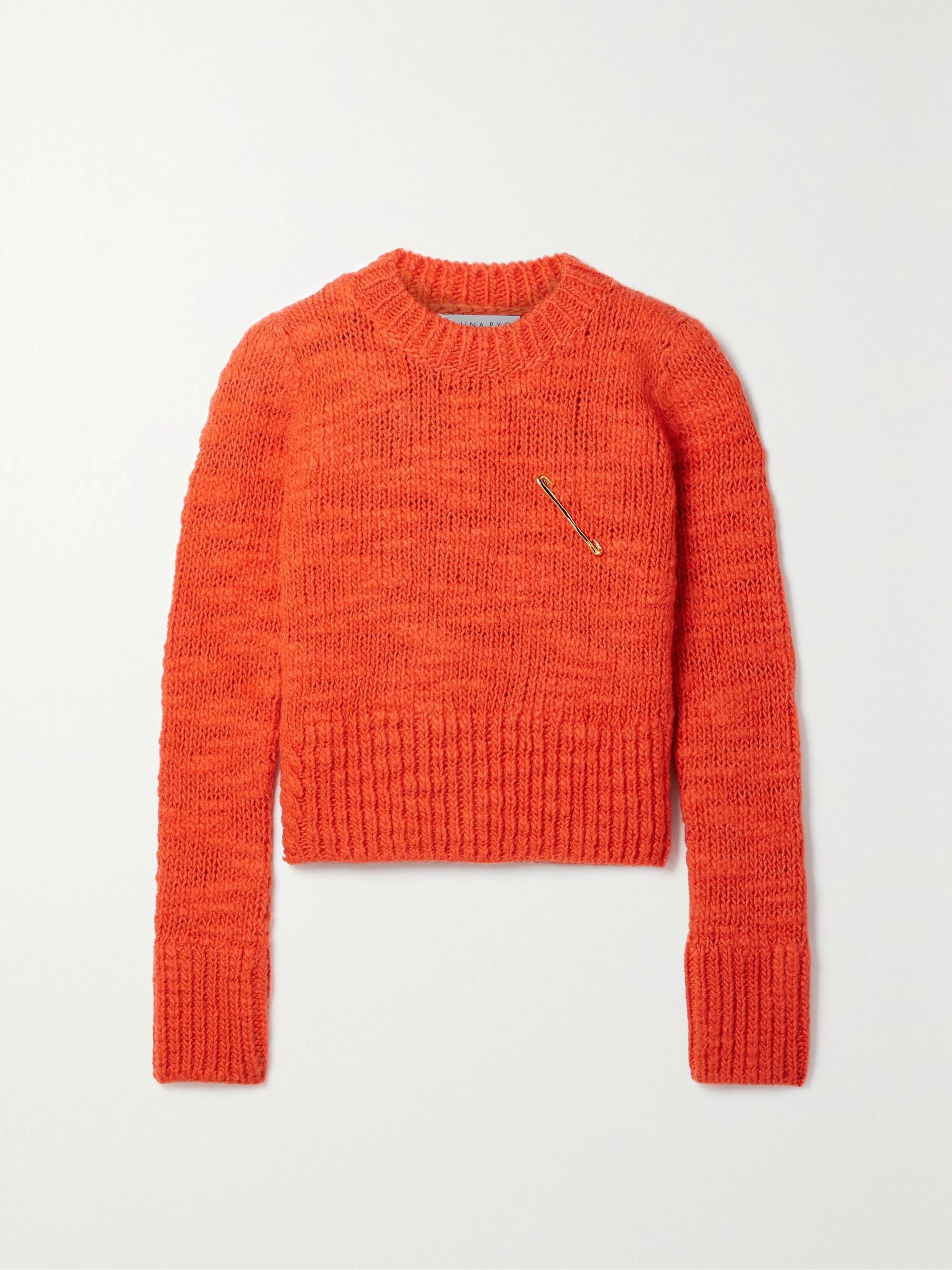 REJINA PYO Wayne embellished wool cropped knit sweater in bright orange comes in a cropped fit with chunky rib knit at the hem, cuff and neck. It has a round neck and removable gold safety pin brooch detail attached at the chest. 